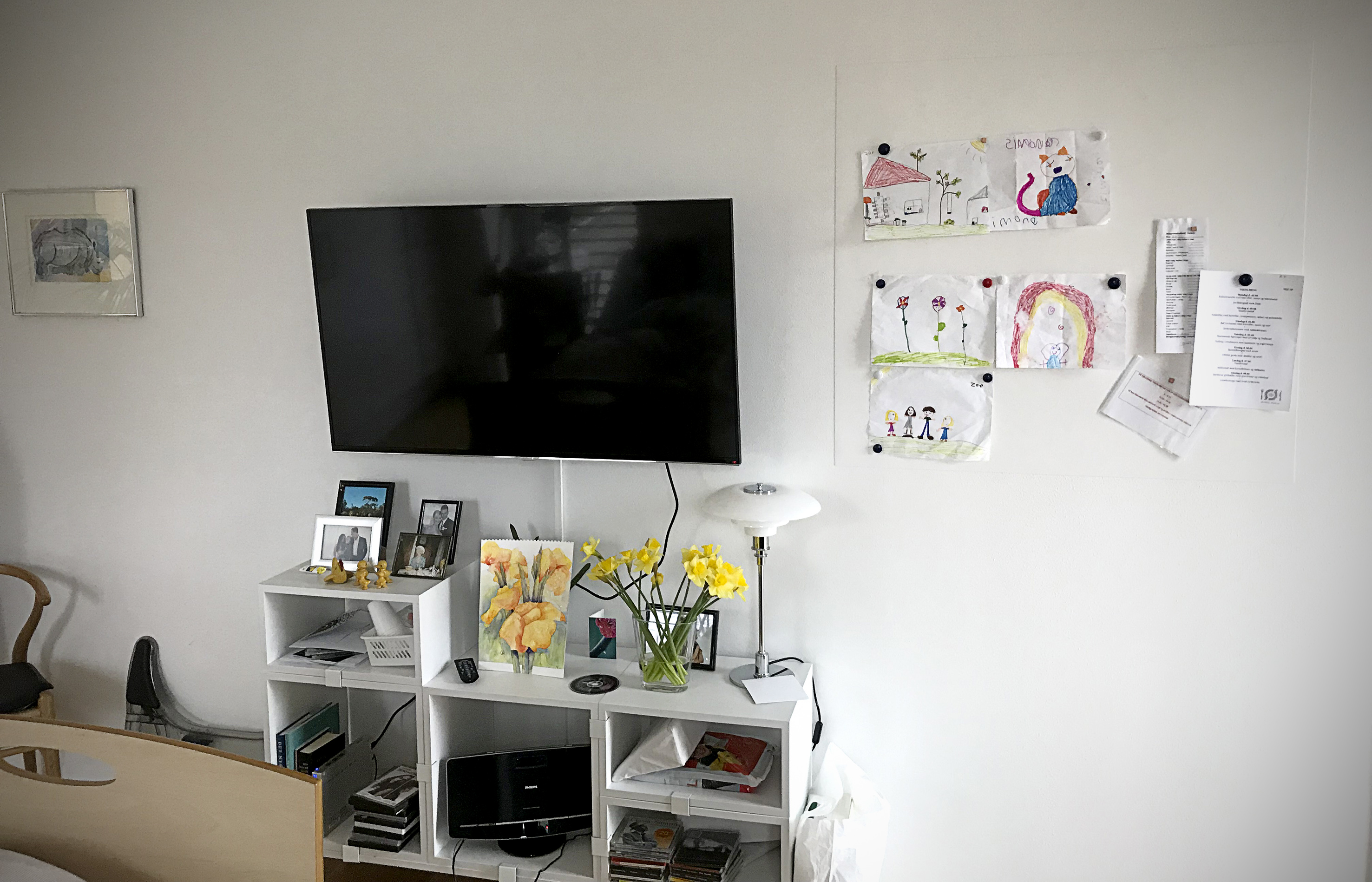 Billedet viser en væg med et fjernsyn og børnetegninger hængt op ved siden af.