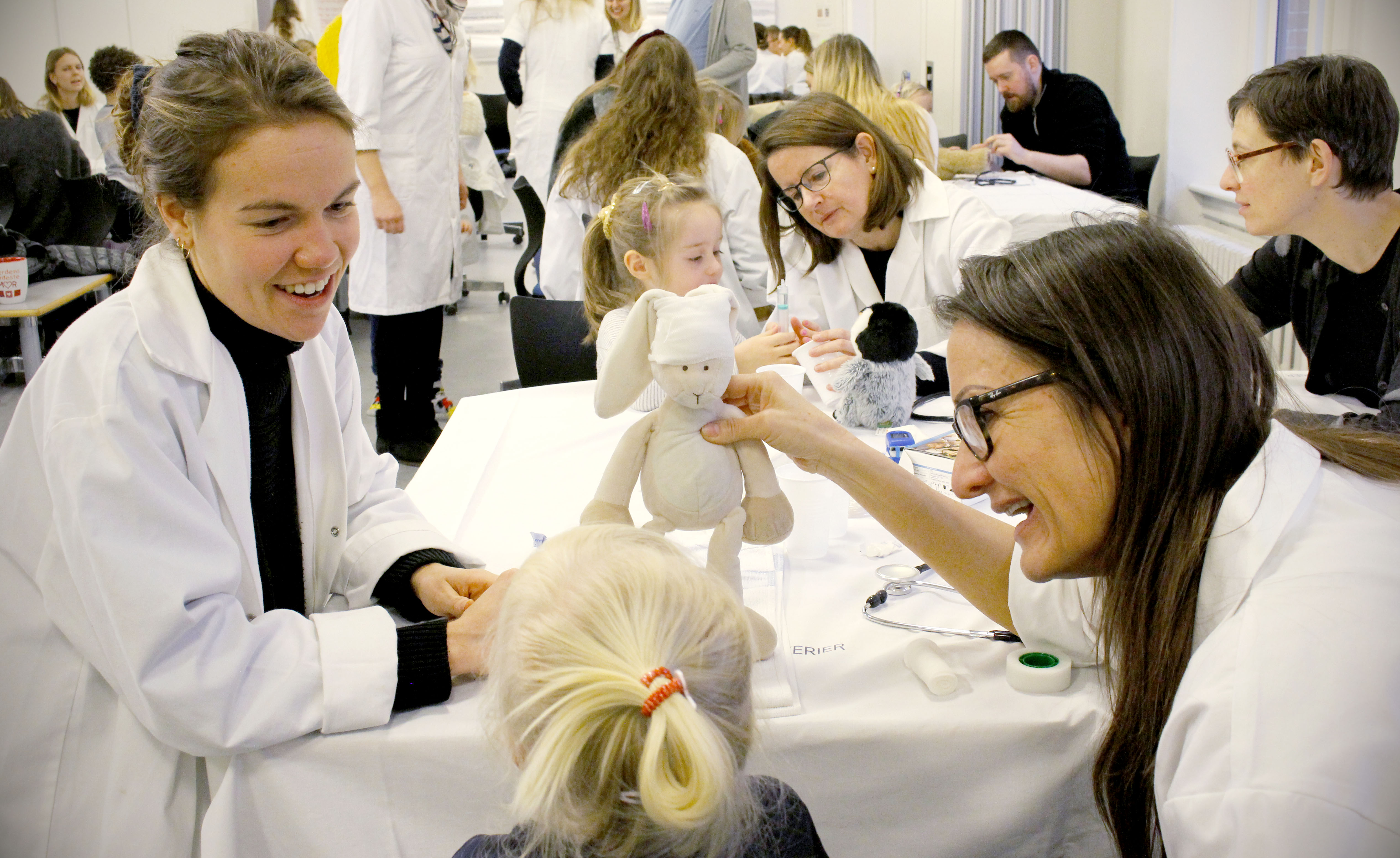 To sygeplejerskestuderende sidder med en lille pige og giver hendes bamse en bandage på.