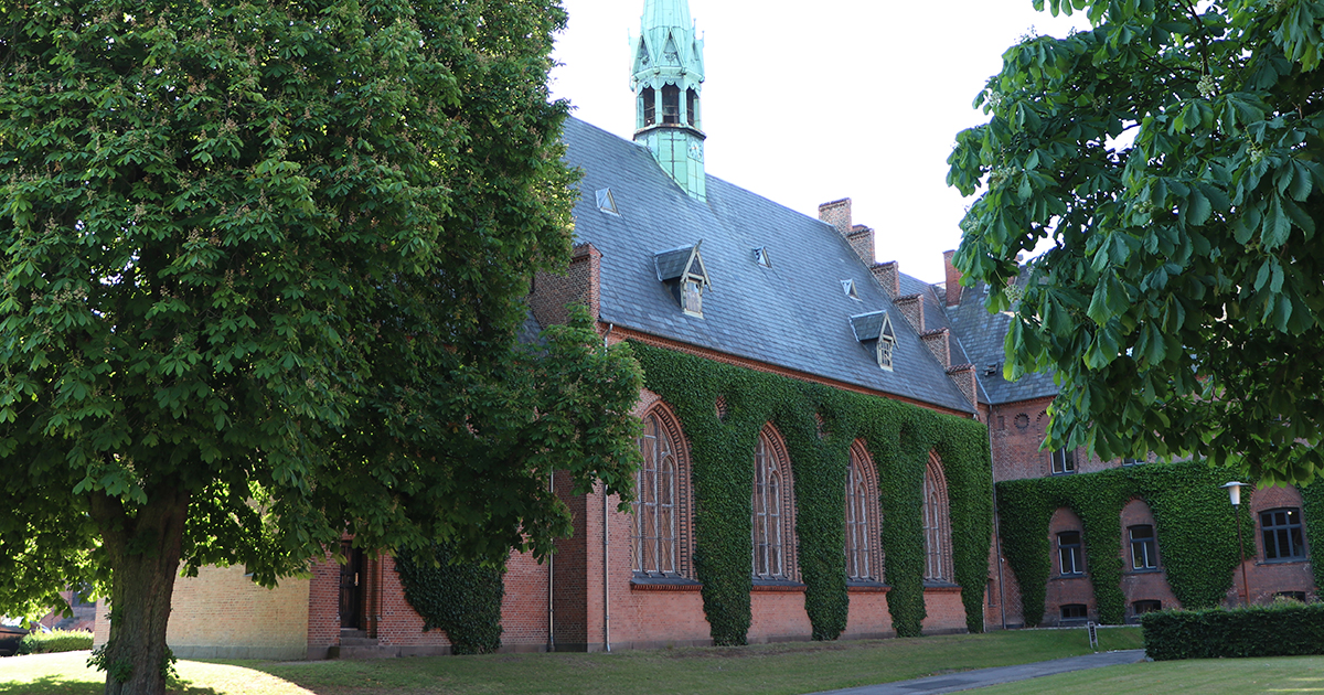 Foto af en kirkebygning i røde mursten med grønne træer på begge sider.