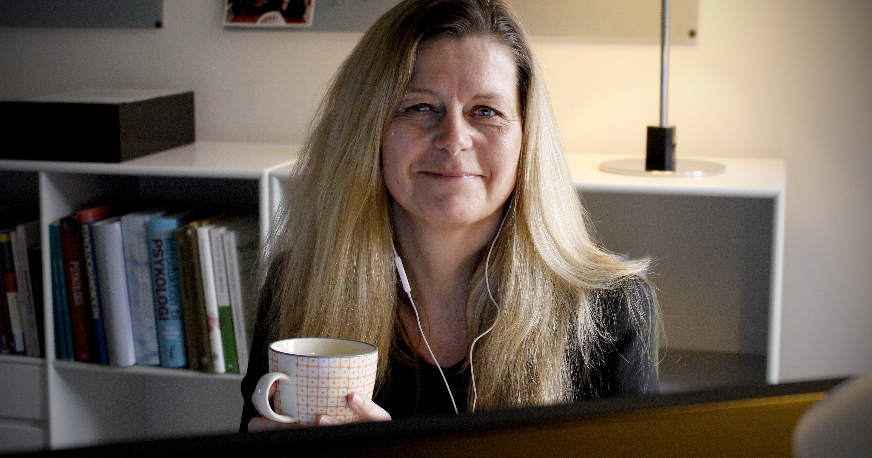 Foto af Rikke Vinter Hedensted ved en computerskærm med en kaffekop i hånden og et headset i øret.