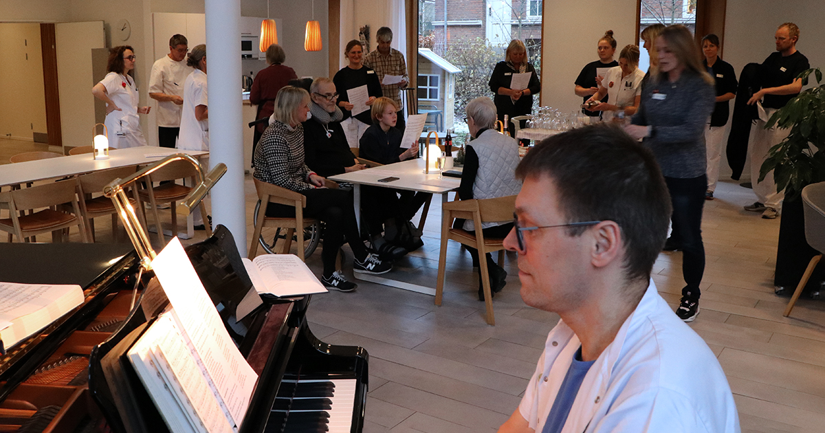 Billedet viser et antal mennesker, som spiser og drikker, mens en mand i hvidt tøj spiller klaver.
